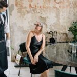 A stylish woman in a black dress interacts with a waiter wearing a face mask in a trendy indoor café setting.