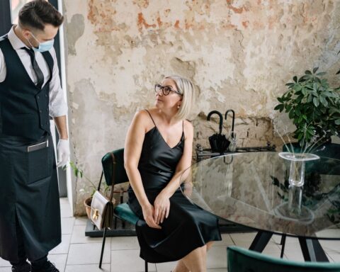 A stylish woman in a black dress interacts with a waiter wearing a face mask in a trendy indoor café setting.