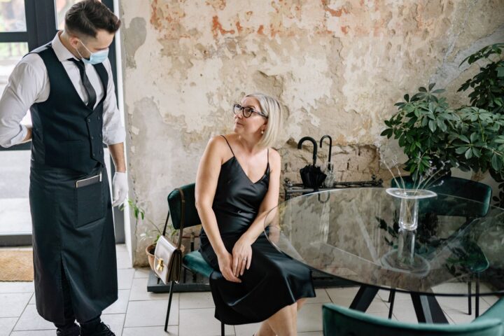 A stylish woman in a black dress interacts with a waiter wearing a face mask in a trendy indoor café setting.