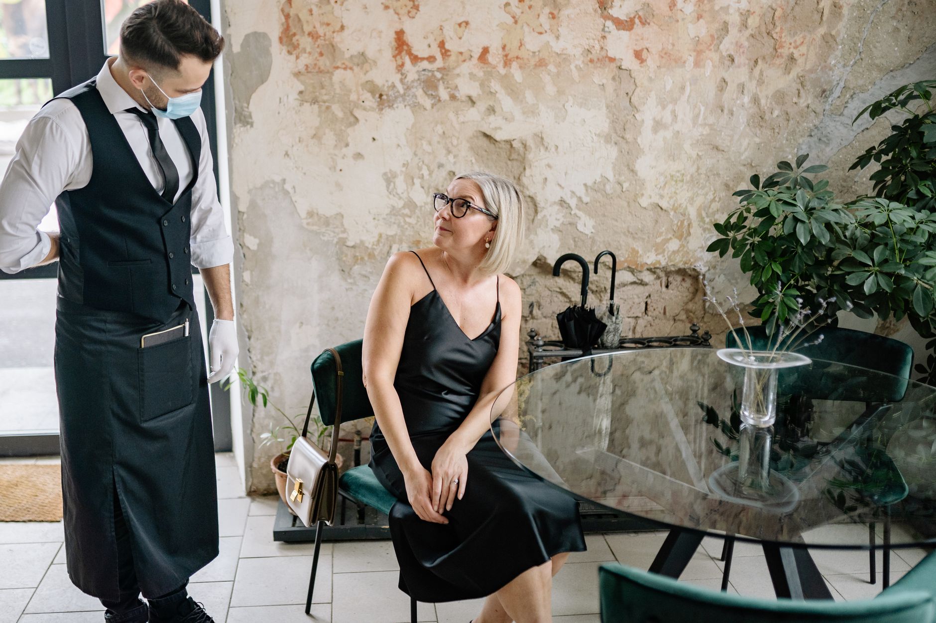 A stylish woman in a black dress interacts with a waiter wearing a face mask in a trendy indoor café setting.