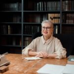 Senior woman sitting at desk in library, smiling at camera, with books and typewriter.
