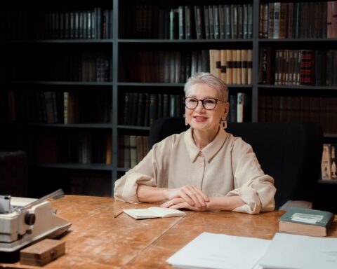 Senior woman sitting at desk in library, smiling at camera, with books and typewriter.