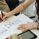 Fashion designer sketching dress ideas at a desk with fabric and sketches.