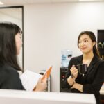 Two businesswomen conversing in an office setting, holding a coffee mug.