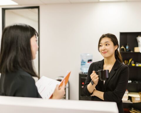 Two businesswomen conversing in an office setting, holding a coffee mug.