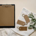 A minimalist flat lay of brown clipboard and tags with green leaves on white cloth.