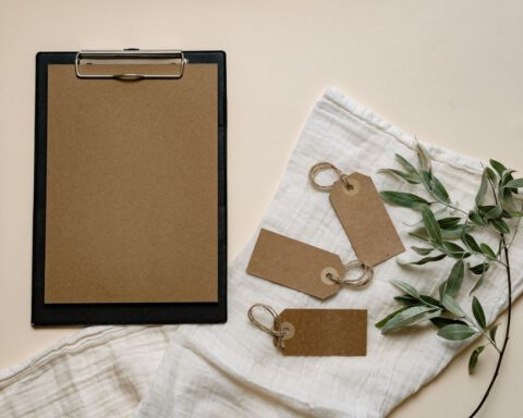 A minimalist flat lay of brown clipboard and tags with green leaves on white cloth.