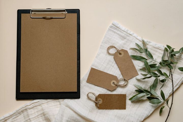 A minimalist flat lay of brown clipboard and tags with green leaves on white cloth.