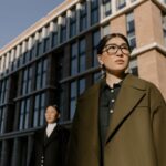 Two professional women in elegant coats stand confidently outside a contemporary urban office building.