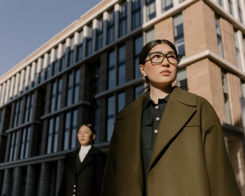 Two professional women in elegant coats stand confidently outside a contemporary urban office building.
