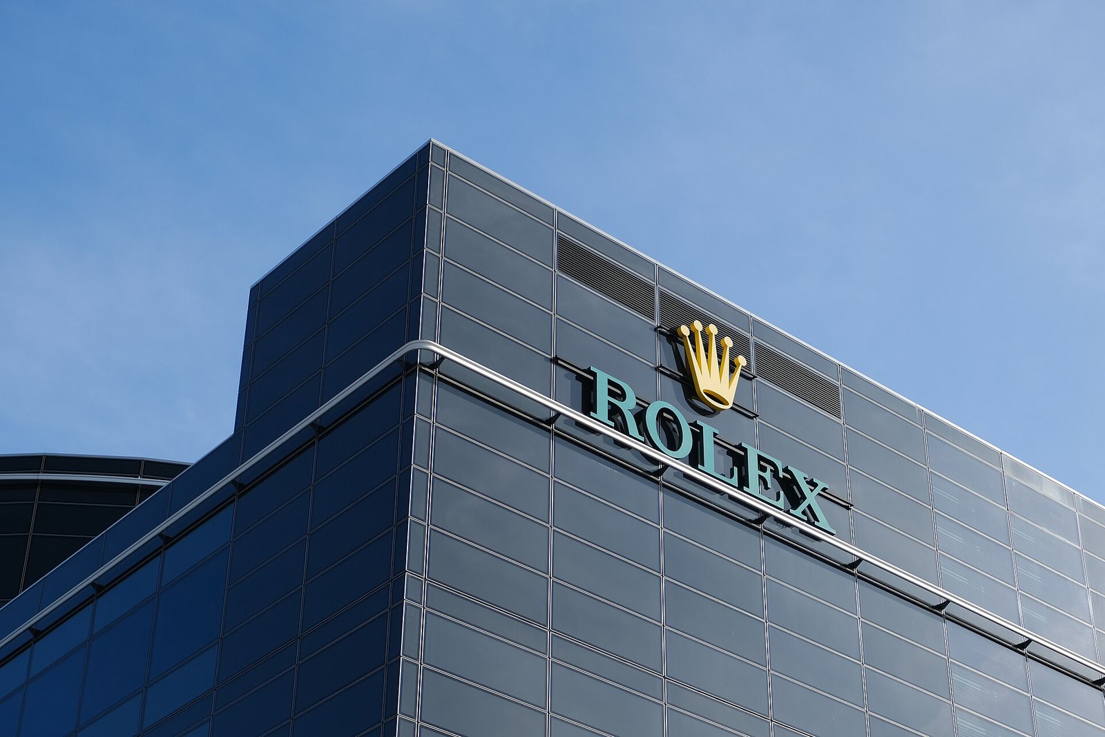 Rolex crown logo and name on the dark glass facade of the Rolex headquarters building in Geneva against a blue sky
