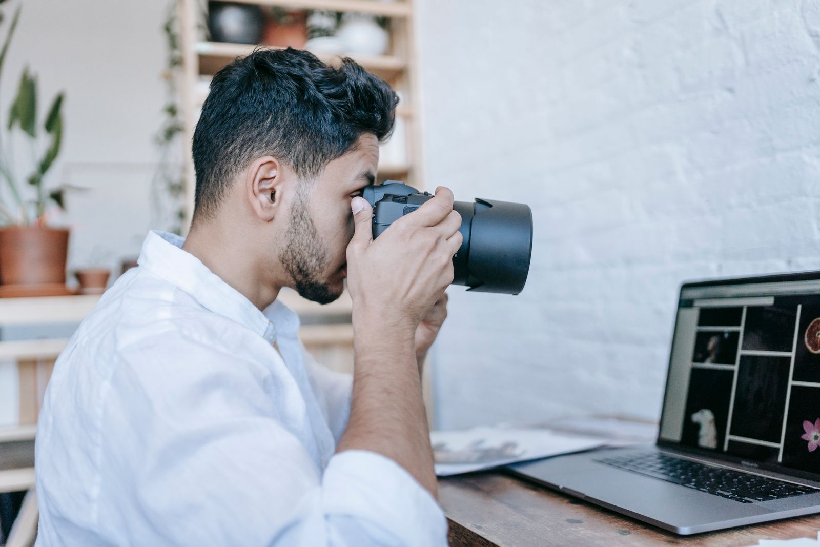 Photographer looking through a camera beside a laptop showing a grid of images