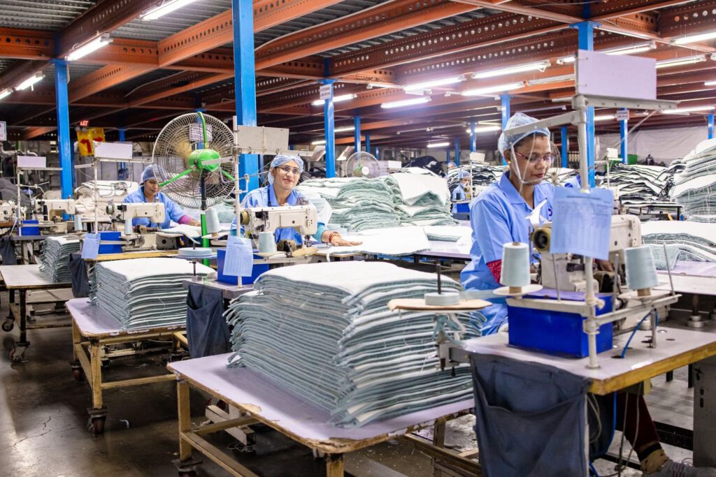Garment workers in blue uniforms and hairnets sewing at industrial machines beside stacks of fabric