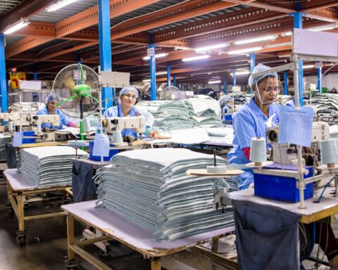 Garment workers in blue uniforms and hairnets sewing at industrial machines beside stacks of fabric
