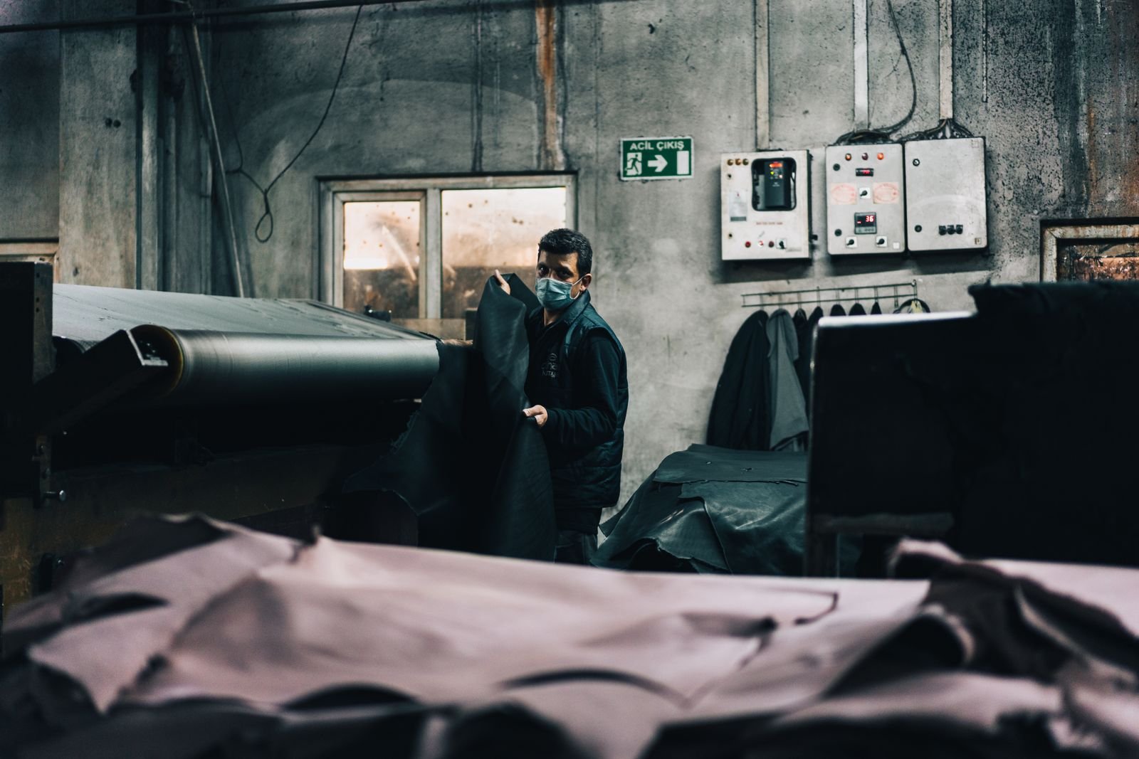 Tannery worker in a face mask lifting dark leather sheets from a roller machine with hides stacked in the foreground