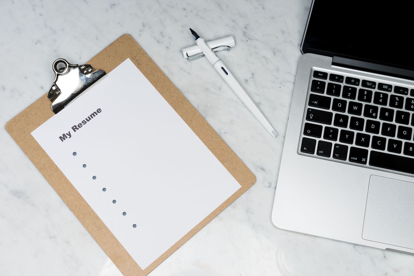 Clipboard holding a blank resume beside a laptop and a white fountain pen on a marble desk