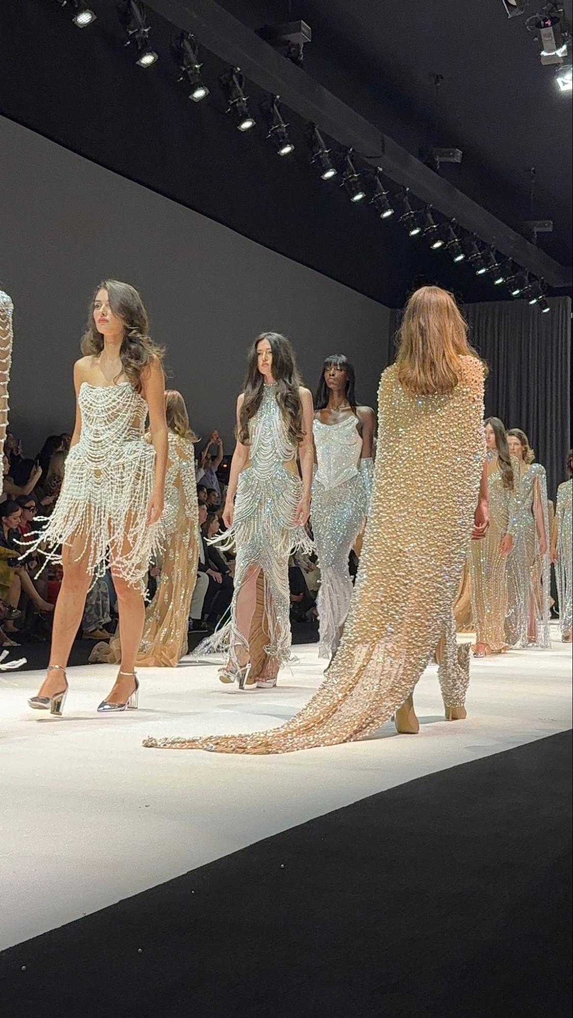 Models in crystal and pearl embellished gowns and minidresses on the runway at Dubai Fashion Week, one wearing a long beaded cape