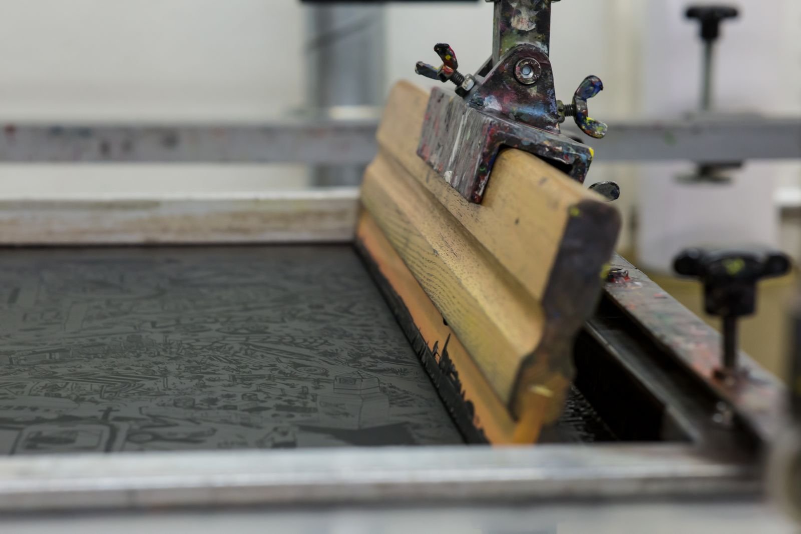 Wooden squeegee clamped over an inked screen on a screen printing press