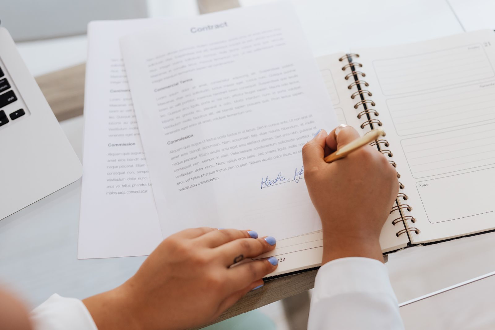 Person signing a printed contract with a commission clause next to a laptop and planner