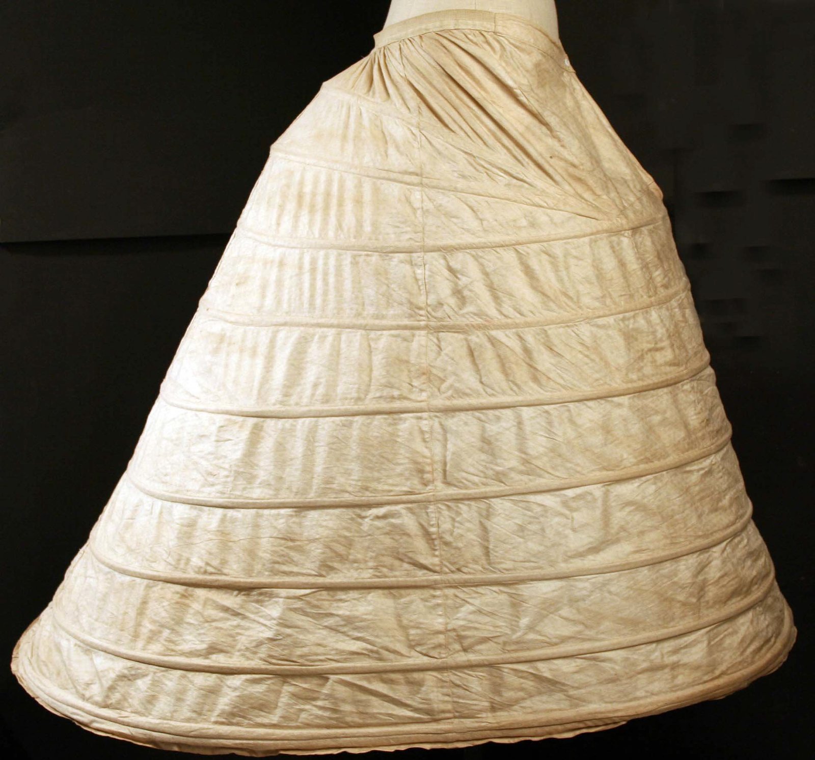 British cage crinoline from about 1862 with fabric-covered steel hoops widening towards the hem, on a dark background