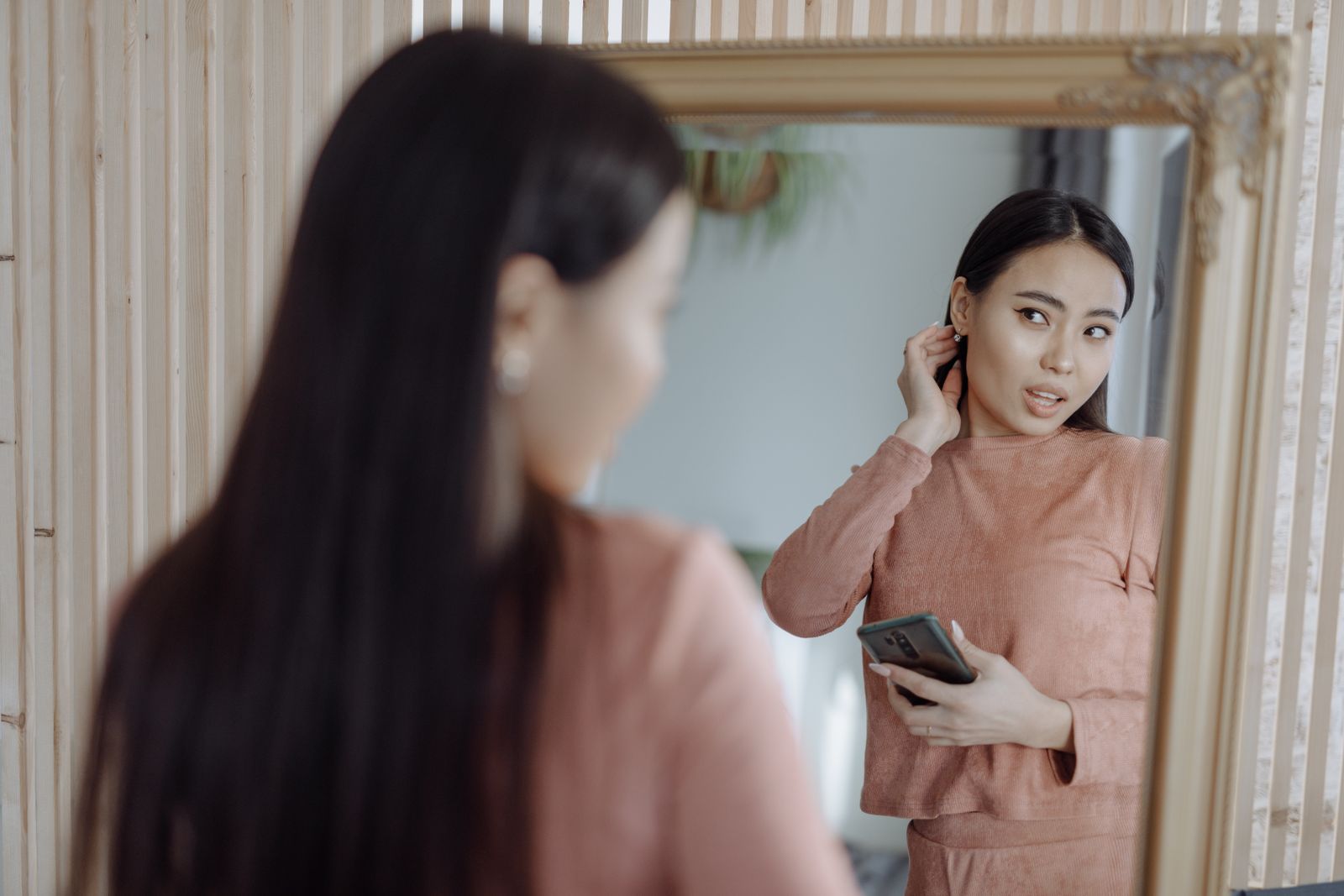 Woman holding a smartphone and checking her face and earring in a large mirror